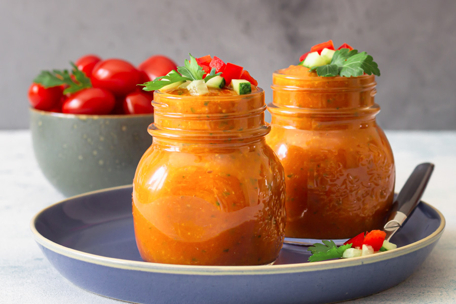 recette de gaspacho Image d'ambiance : Deux bocaux à couvercle vissé remplis de gaspacho rouge disposés sur une assiette bleue, en arrière-plan un bol de tomates